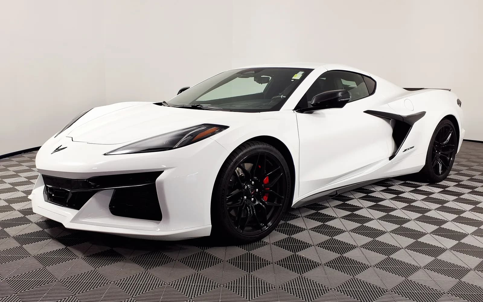 Chevrolet Corvette Z06 Coupé (C8) blanco en tres cuartos frontal izquierdo dentro del salón de un concesionario. Se distinguen la arquitectura de motor central con contrafuertes traseros, los guardafangos ensanchados, la gran toma de aire lateral negra, el emblema Z06 en el panel trasero y los rines negros con pinzas de freno rojas. Imagen referencial del modelo, no de la unidad en venta.