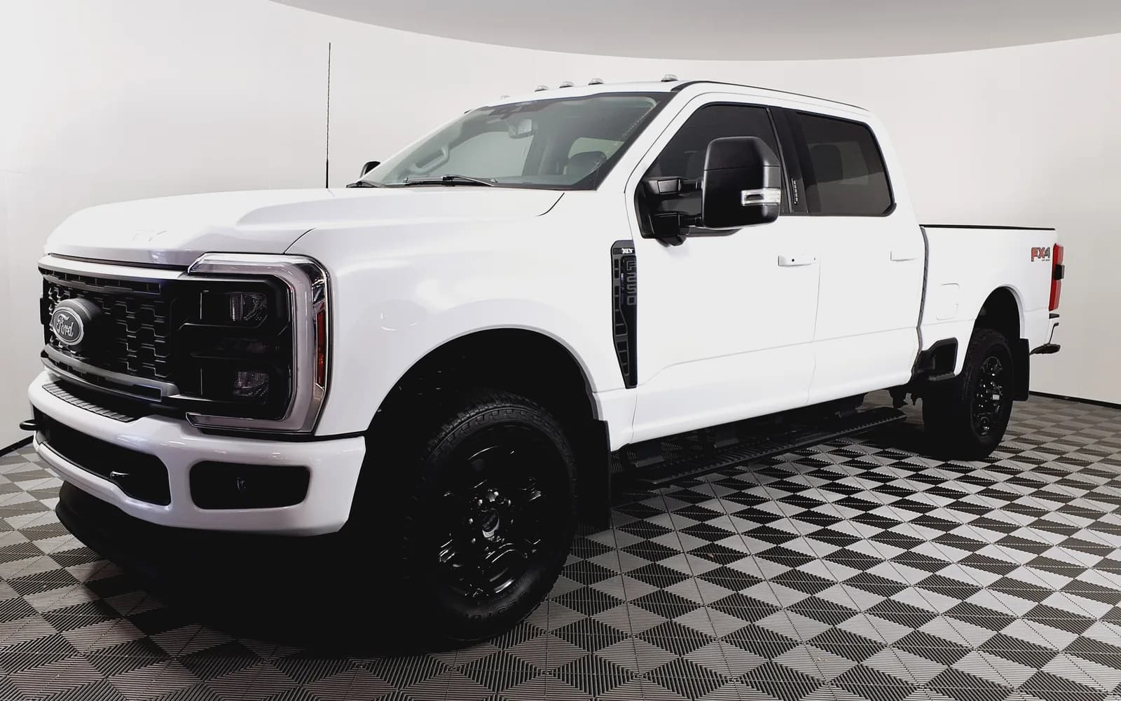 Ford F-250 Super Duty XLT blanco de cabina doble, generación P708 (2023-2026), en tres cuartos frontal izquierdo dentro de una cabina de fotografía de concesionario con piso ajedrezado. Se distinguen la parrilla de dos barras, los faros LED con firma en doble C, los espejos de remolque y el decal FX4 en el platón. Imagen referencial del modelo, no de la unidad en venta.