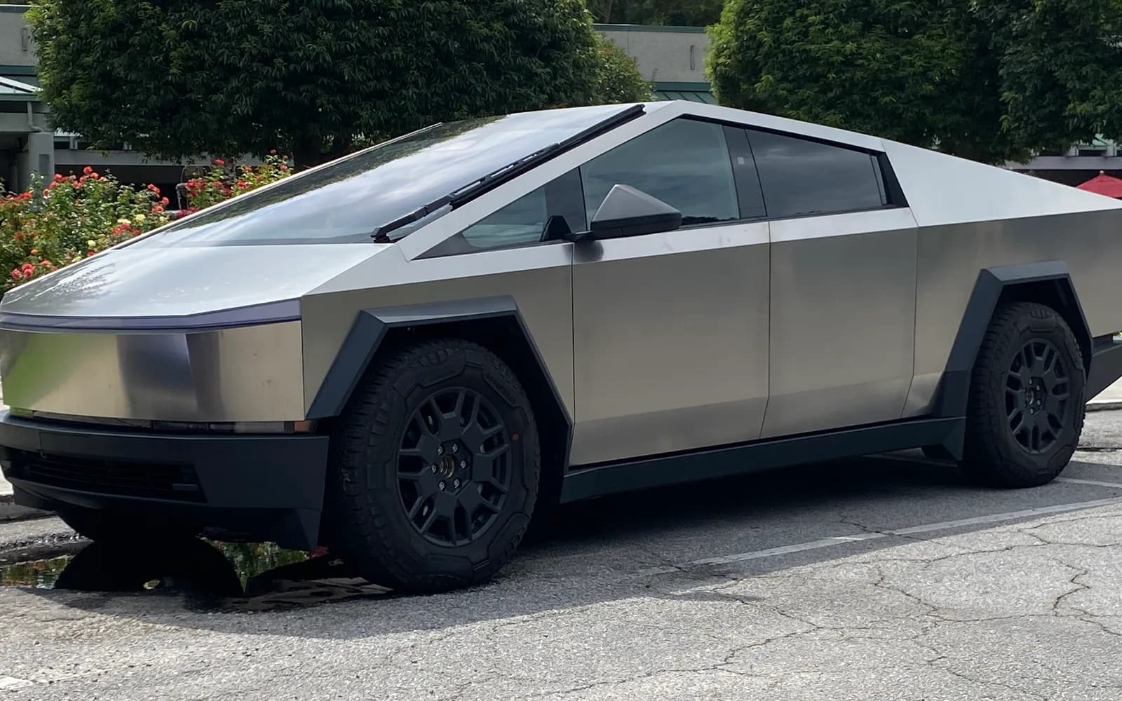 Tesla Cybertruck en acero inoxidable sin pintar, vista frontal izquierda escorada hacia el perfil, estacionado sobre pavimento con setos y rosales de fondo. Se distinguen la carrocería en cuña de paneles planos, el parabrisas de una sola pieza, la franja LED delantera continua y los guardafangos y llantas negros. Imagen referencial del modelo, no de la unidad en venta.
